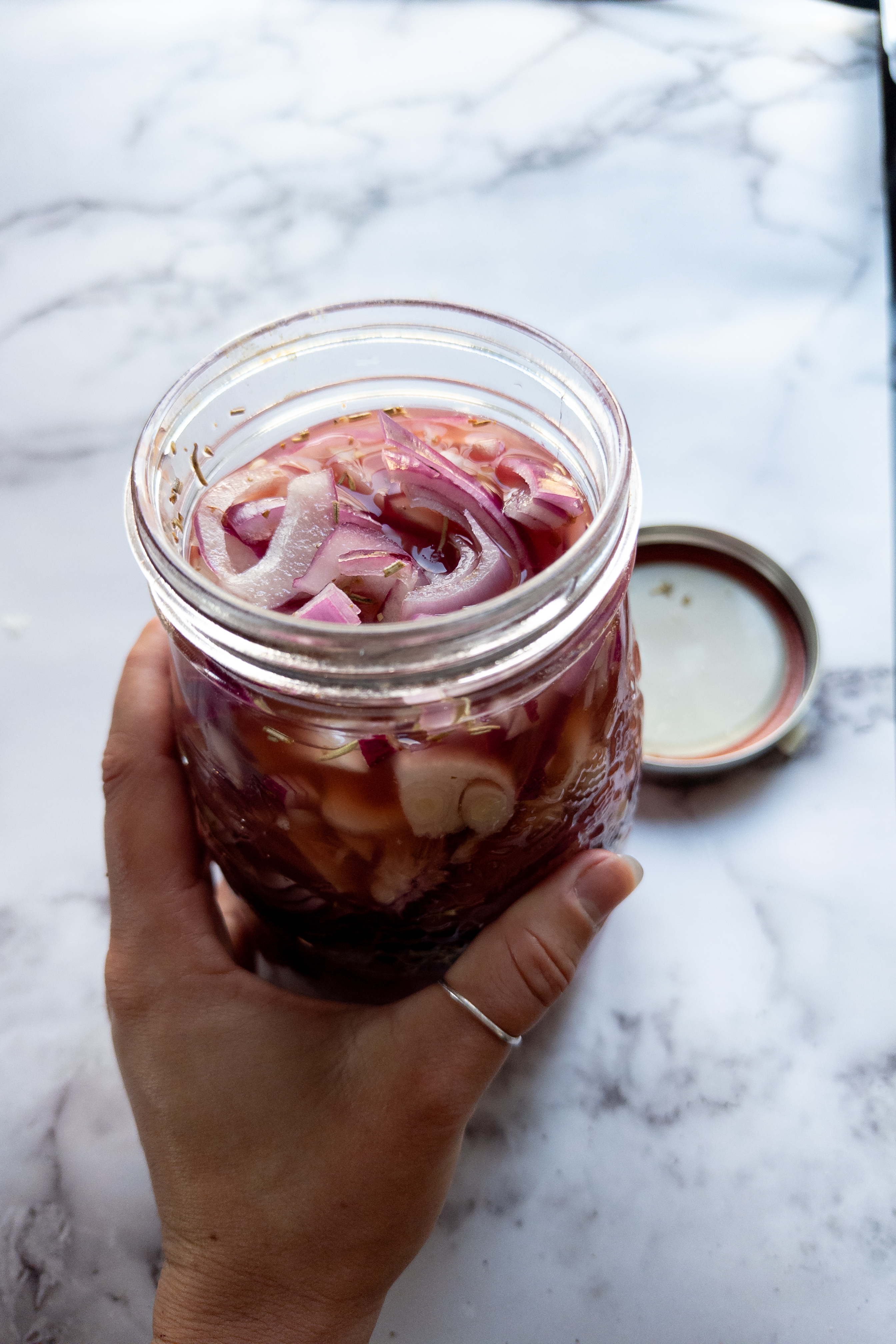 red onions cut in slices and placed in a jar with herbs and garlic on top and hand holding jar