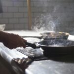 A chef cooks with frying pans on an electric stove in a steamy kitchen.