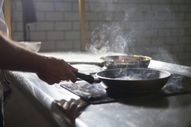 A chef cooks with frying pans on an electric stove in a steamy kitchen.